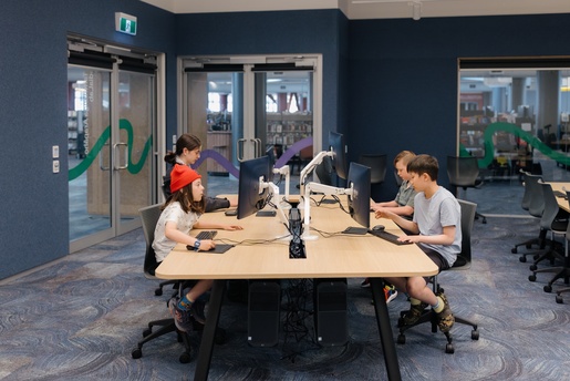 Four young people sit at a large table in a modern library or computer lab, each working on desktop computers, with blue walls, glass doors, and bookshelves in the background