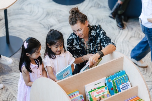 A person is helping two young children choose books at a curved wooden bookshelf in a bright library. The children hold picture books while standing near shelves filled with colorful titles, with patterned flooring visible below.