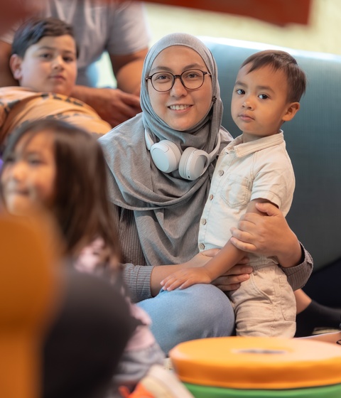 A person stands indoors looking up at three large, brightly colored vertical digital artworks depicting stylized figures in vibrant reds, purples, and golds. The illuminated panels dominate the space with bold patterns and warm light.