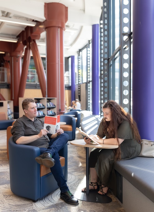 Two people read in a bright modern library space, one in a blue armchair holding a book and the other on a cushioned bench, with tall patterned windows, red columns, and bookshelves in the background.