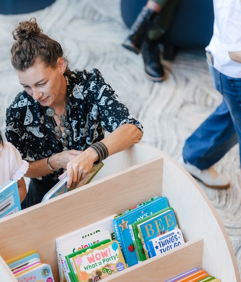A person is helping two young children choose books at a curved wooden bookshelf in a bright library. The children hold picture books while standing near shelves filled with colorful titles, with patterned flooring visible below.