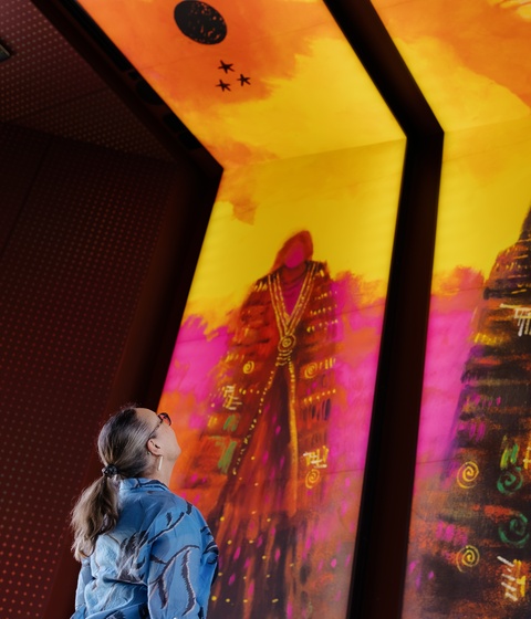 A person stands indoors looking up at three large, brightly colored vertical digital artworks depicting stylized figures in vibrant reds, purples, and golds. The illuminated panels dominate the space with bold patterns and warm light.
