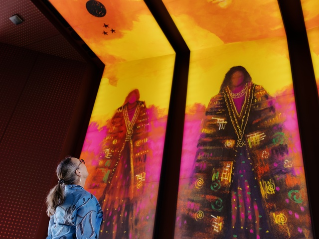 A person stands indoors looking up at three large, brightly colored vertical digital artworks depicting stylized figures in vibrant reds, purples, and golds. The illuminated panels dominate the space with bold patterns and warm light.