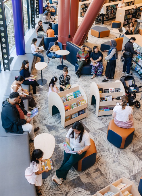 People read, browse shelves, and sit at tables in a bright, modern library filled with curved bookshelves, colorful books, large windows, and cushioned seating.