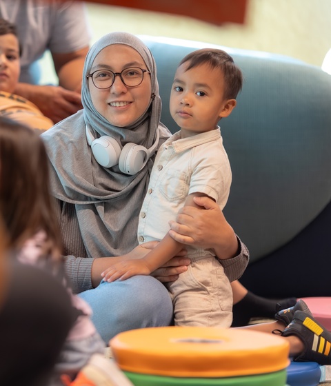 A person stands indoors looking up at three large, brightly colored vertical digital artworks depicting stylized figures in vibrant reds, purples, and golds. The illuminated panels dominate the space with bold patterns and warm light.