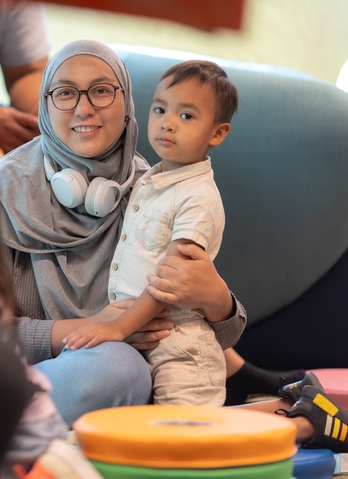 A person stands indoors looking up at three large, brightly colored vertical digital artworks depicting stylized figures in vibrant reds, purples, and golds. The illuminated panels dominate the space with bold patterns and warm light.