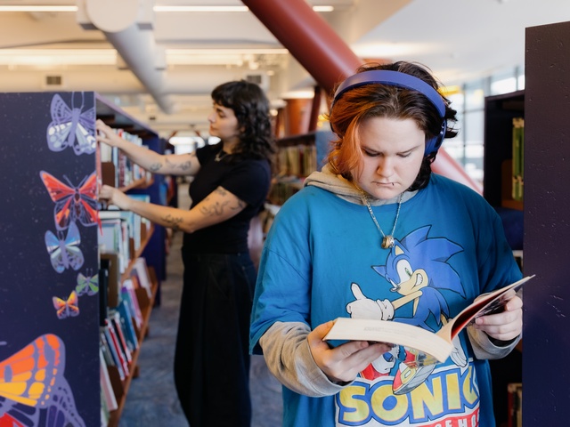 A person wearing a blue Sonic‑the‑Hedgehog shirt, gray hoodie, and blue headphones reads a book indoors, with warm lighting and shelves in the background.