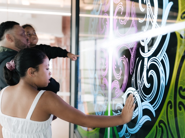 A child touches a large interactive digital display showing swirling neon colors, while an adult behind them holds another child who watches the screen. The scene appears in a modern, well‑lit museum or tech exhibit