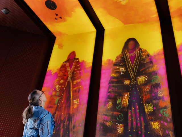 A person stands indoors looking up at three large, brightly colored vertical digital artworks depicting stylized figures in vibrant reds, purples, and golds. The illuminated panels dominate the space with bold patterns and warm light.