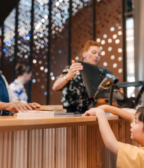 A staff member stands behind a wooden counter assisting a visitor and a child who is reaching toward the counter. Additional people are visible in the background near decorative panels with circular cut‑out patterns.
