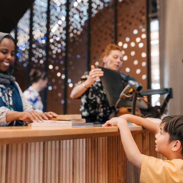 A staff member stands behind a wooden counter assisting a visitor and a child who is reaching toward the counter. Additional people are visible in the background near decorative panels with circular cut‑out patterns.