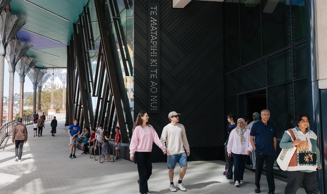People walk and gather outside a modern building with tall patterned columns and a shaded walkway. Some individuals are seated while others stroll near the entrance, where the wall displays the text “TE MATAPIHI KI TE AO NUI.”