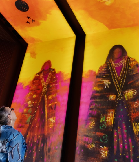 A person stands indoors looking up at three large, brightly colored vertical digital artworks depicting stylized figures in vibrant reds, purples, and golds. The illuminated panels dominate the space with bold patterns and warm light.