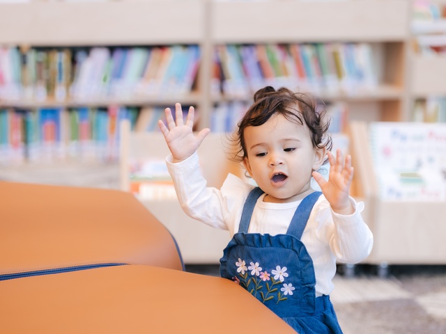 A young child in blue overalls with flower designs raises both hands while standing in front of colorful bookshelves, with orange cushioned seating in the foreground.