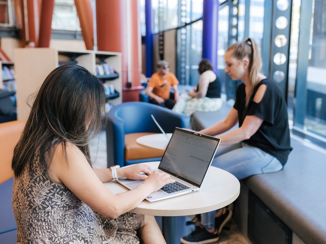 People using laptops and seating areas inside Te Matapihi library, with others studying and relaxing in the background.