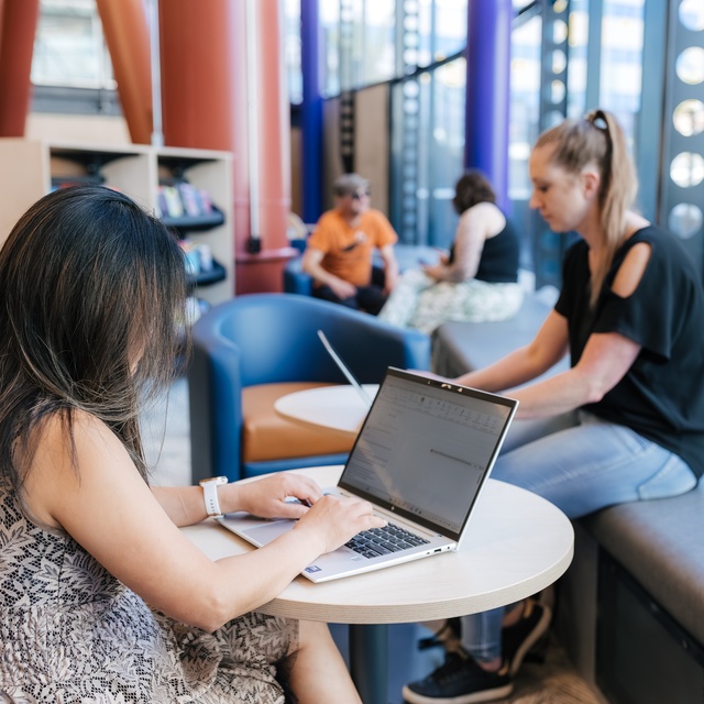 People using laptops and seating areas inside Te Matapihi library, with others studying and relaxing in the background.
