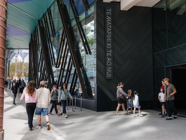 People walk past the entrance of a modern building with tall angled beams and large glass panels. A sign reading “TE MATAPIHI KI TE AO NUI” is mounted beside the doorway as groups and families move along the wide outdoor walkway.