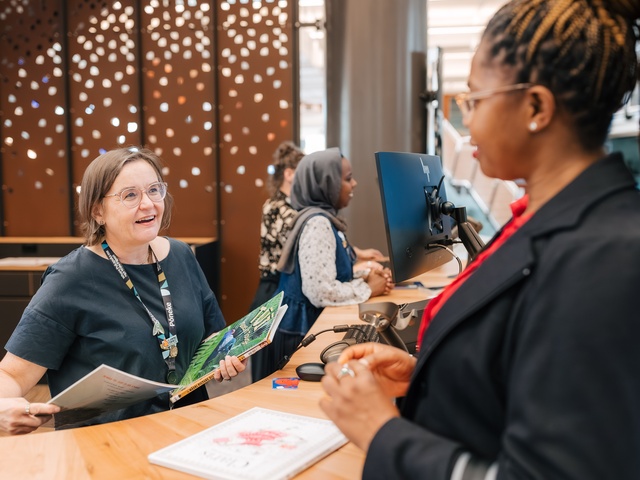 People stand at a service counter in a library or community space. One person holds an open book while others speak with staff behind computer monitors. Decorative panels and books on the counter are visible in the background.