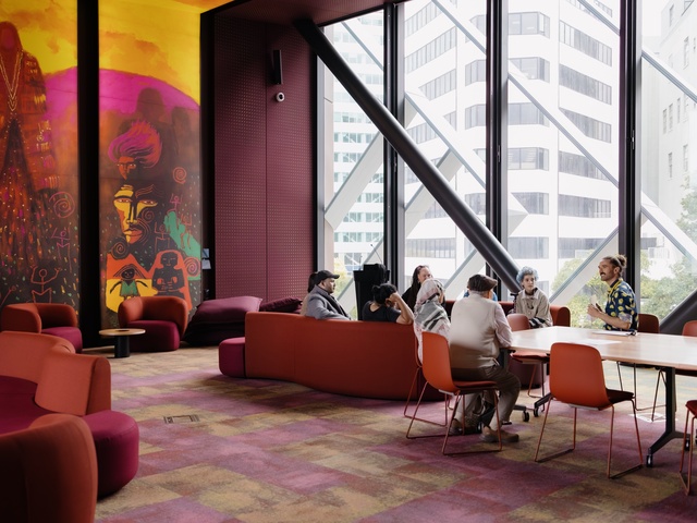 group of people sit around tables and lounge seating in a brightly lit room with large floor‑to‑ceiling windows. Bold, colorful wall art featuring abstract figures fills one side of the space, which is furnished with red and orange chairs and couches.
