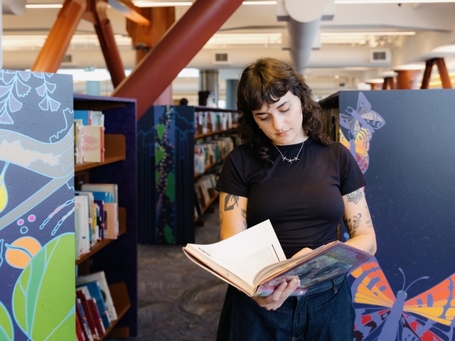 A person stands in a library aisle reading an open book. Colorful panels with illustrated designs frame the shelves, and rows of books extend into the background beneath exposed beams and bright overhead lighting.