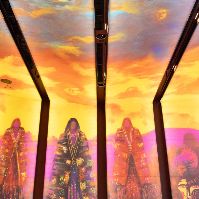 A vibrant ceiling mural shows two large ethereal faces in blue clouds above three robed figures in warm sunset colors, divided by structural beams. The artwork blends spiritual imagery with bold, vivid hues.
