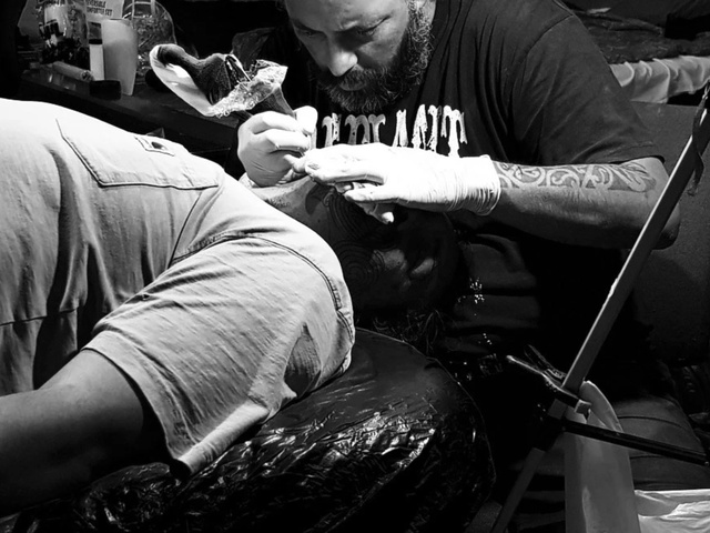 A tattoo artist wearing gloves works on the back of a person lying on a padded surface. The scene is in a dimly lit studio with equipment, artwork, and supplies visible in the background.
