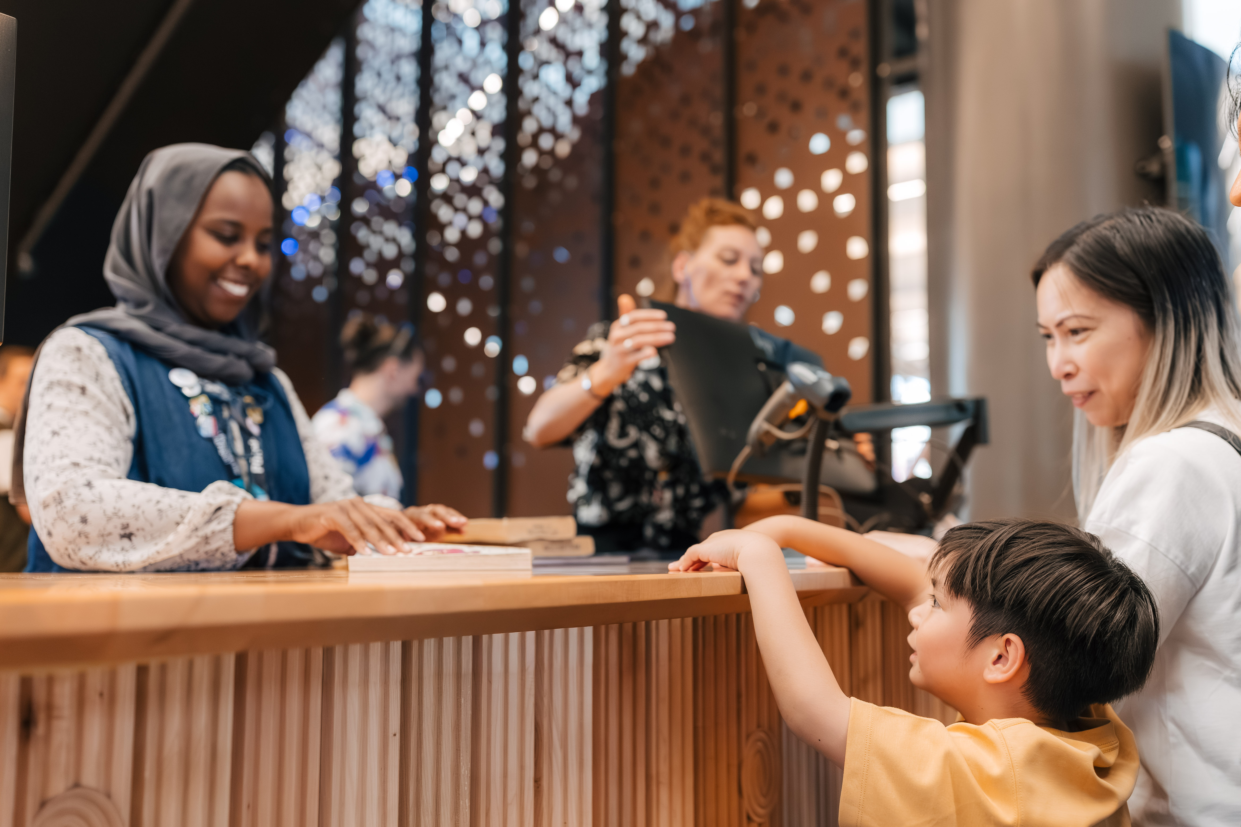 A staff member stands behind a wooden counter assisting a visitor and a child who is reaching toward the counter. Additional people are visible in the background near decorative panels with circular cut‑out patterns.